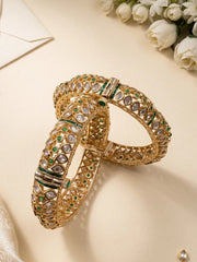An ornate gold-plated uncut polki bangle bracelet decorated with polki stones and small green stones, on a light beige surface with white roses in the background.
