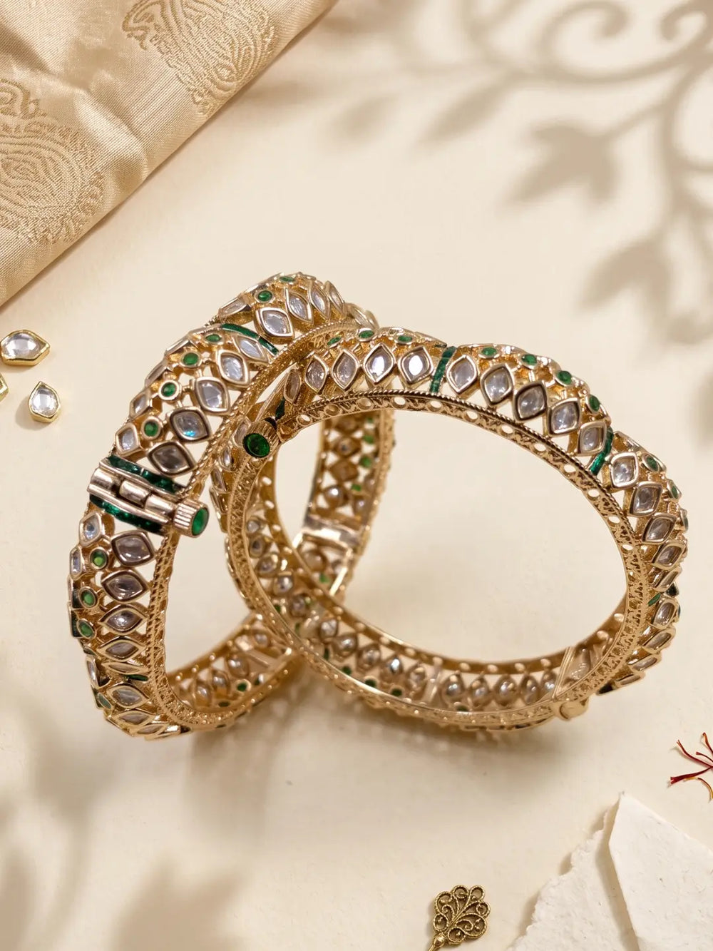 Two ornate gold-toned polki kundan bangles decorated with clear polki stones and green enamel accents, arranged on a cream fabric background with decorative props, perfect for wedding and bridal wear 