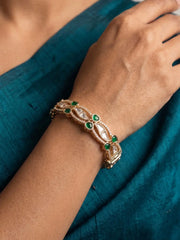 A close-up of a gold-toned traditional Indian bangle bracelet with green stones and white inlay, worn on a person's wrist against a teal fabric background.