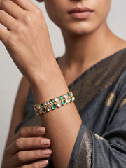 A close-up of a woman wearing a gold-plated bracelet adorned with green and white stones on her wrist, paired with a dark grey saree.