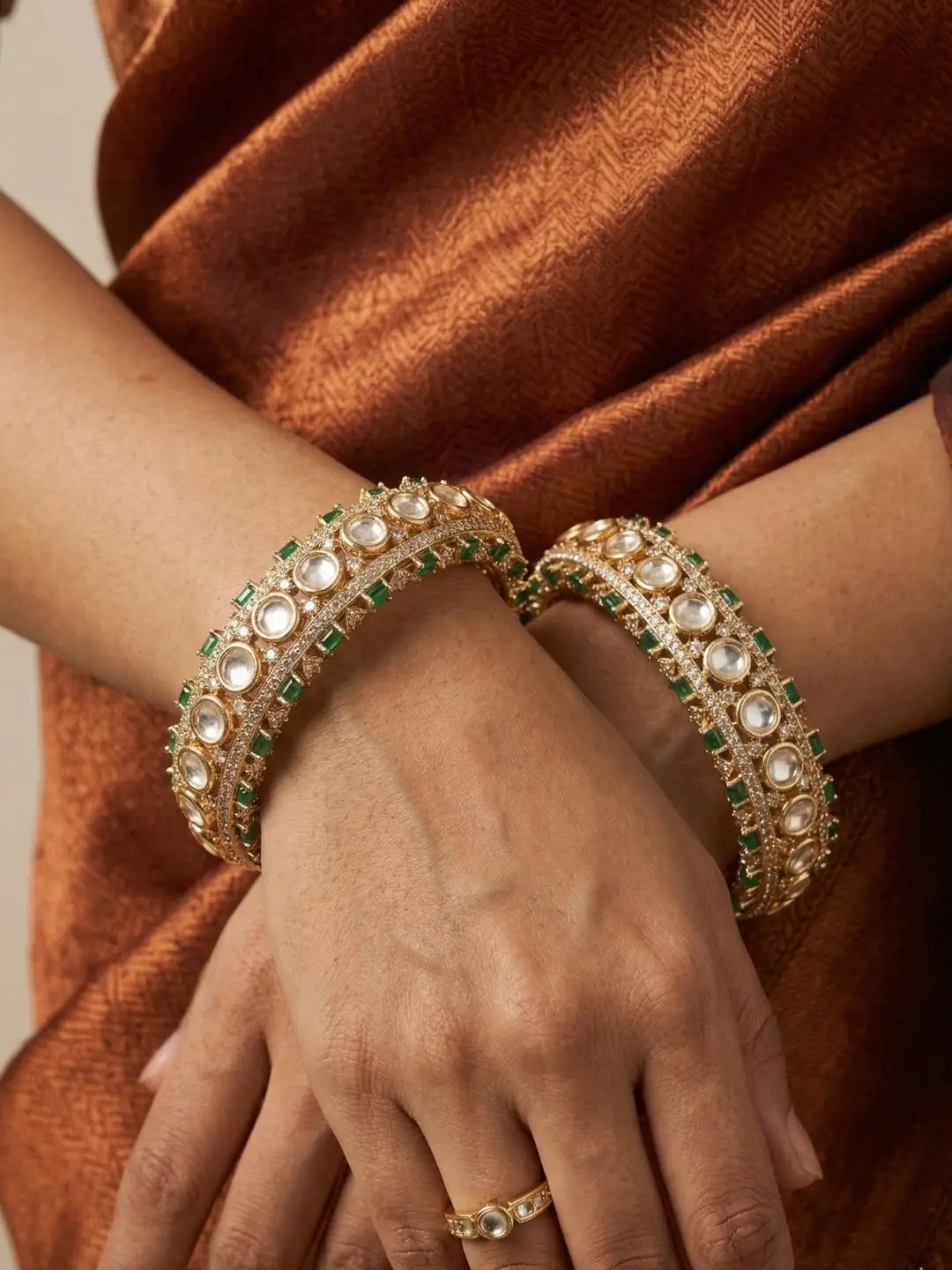 Close-up of a hand wearing two ornate gold bracelets with green stones  and kundan on a brown saree