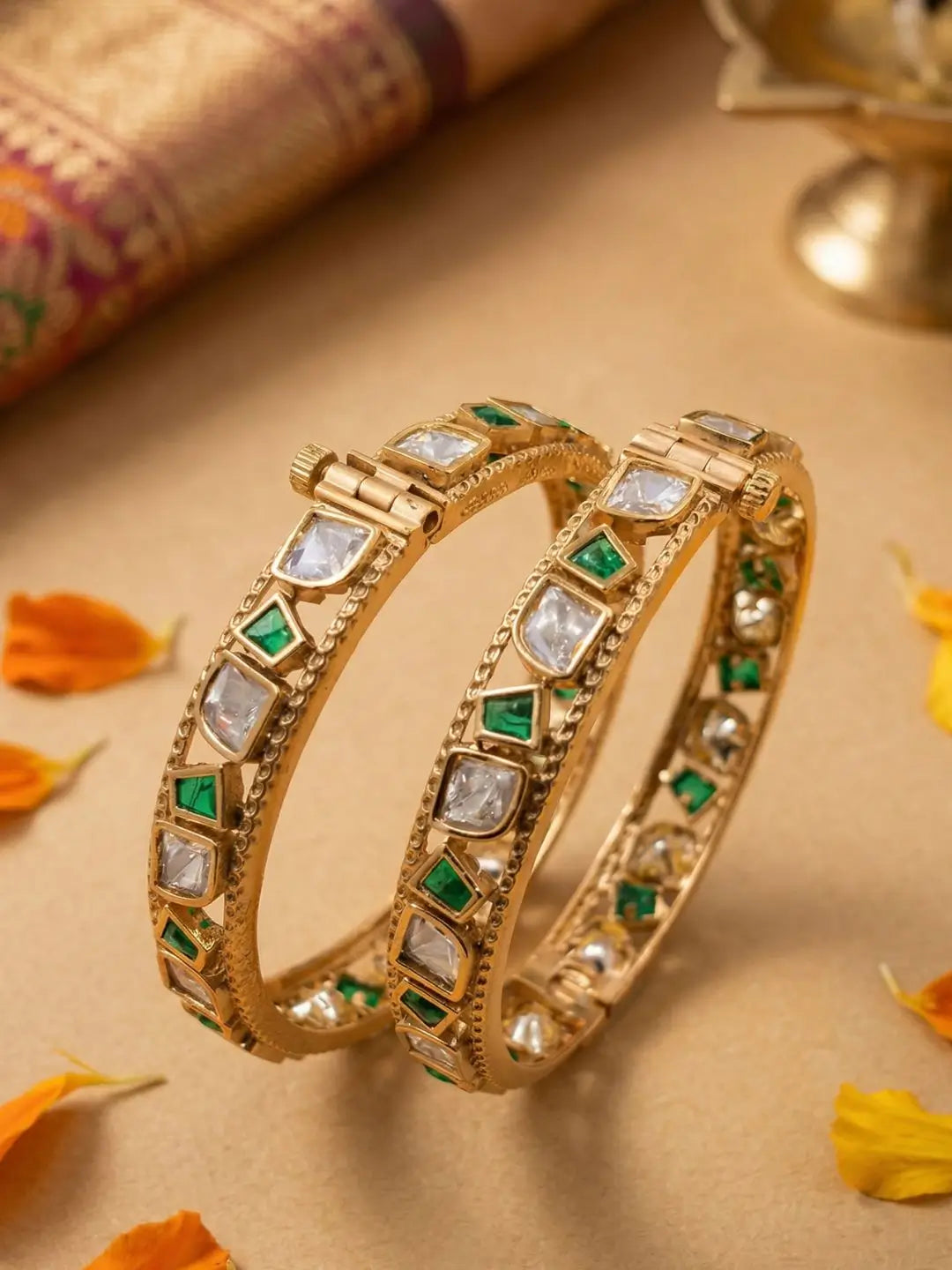Two gold-plated Indian bangles decorated with clear stones and green stones, displayed on a beige surface with decorative flower petals and a traditional fabric roll in the background.