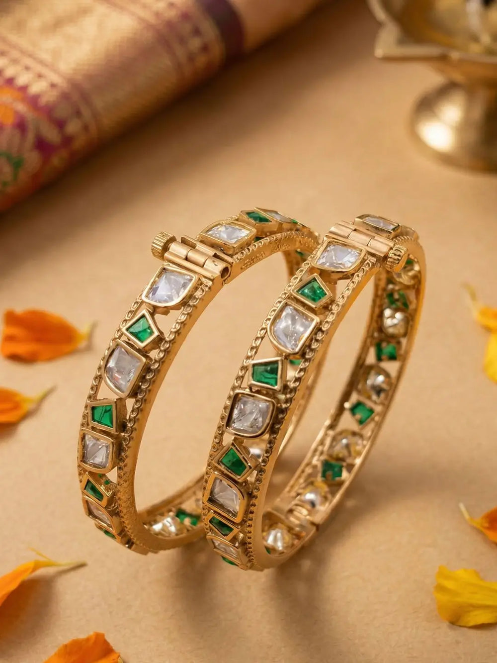 Two gold-plated Indian bangles decorated with clear stones and green stones, displayed on a beige surface with decorative flower petals and a traditional fabric roll in the background.