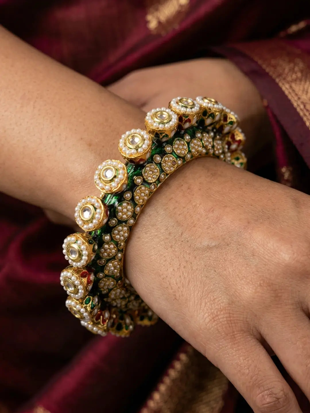 A close-up of a person's wrist and hand wearing an ornate traditional Indian Kundan bangle bracelet. The bracelet is decorated with green enamel, kundan stones, and pearls 