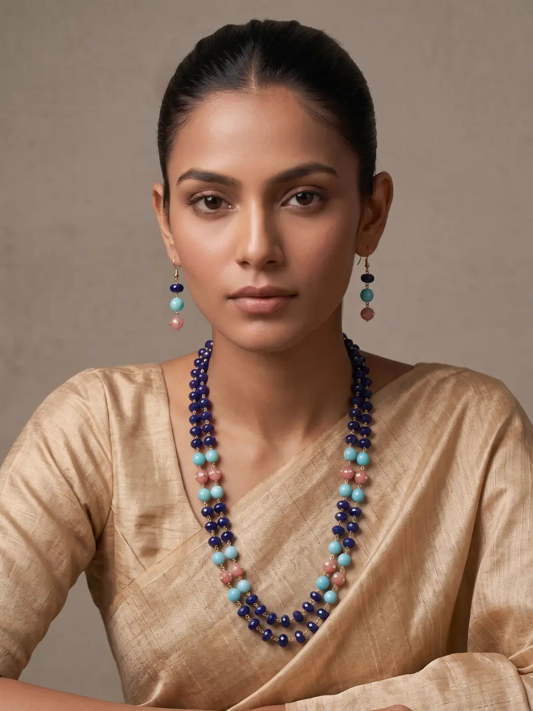 A woman wearing a double-strand beaded necklace and matching drop earrings. The necklace features dark blue beads, light turquoise beads, and dusty pink beads, worn over a beige silk top against a neutral beige background.