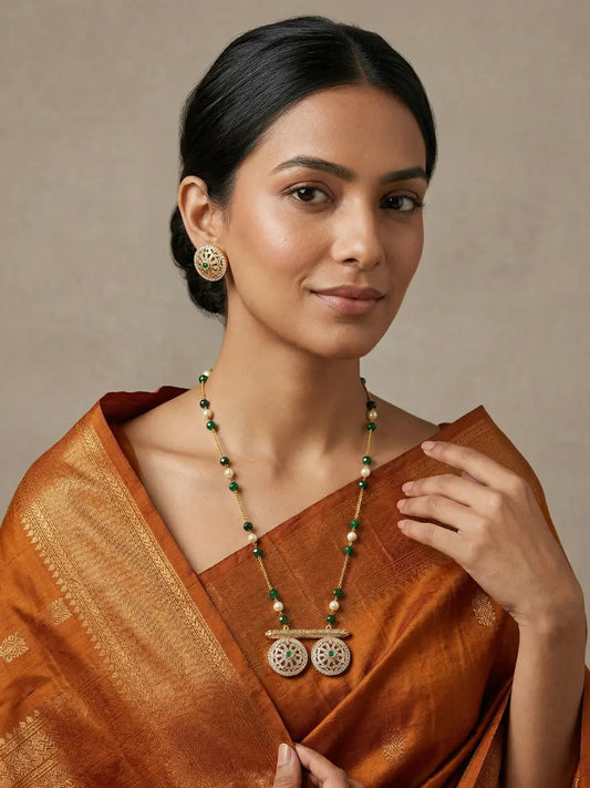 Woman wearing a long green beaded chain necklace with jade beads, faux pearl accents, a circular Polki Meenakari pendant and matching stud earrings.