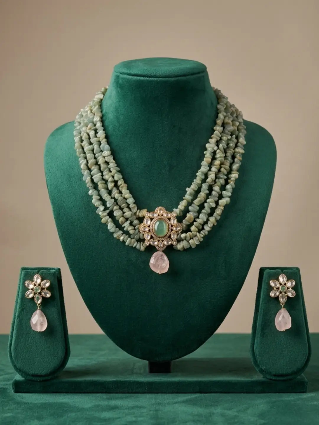A three-piece Indian jewelry set featuring a multi-strand necklace and matching earrings, displayed on a dark green velvet jewelry stand. The jewelry is made of light green gemstone beads with a central gold-toned filigree piece containing a green stone and a pink teardrop pendant.
