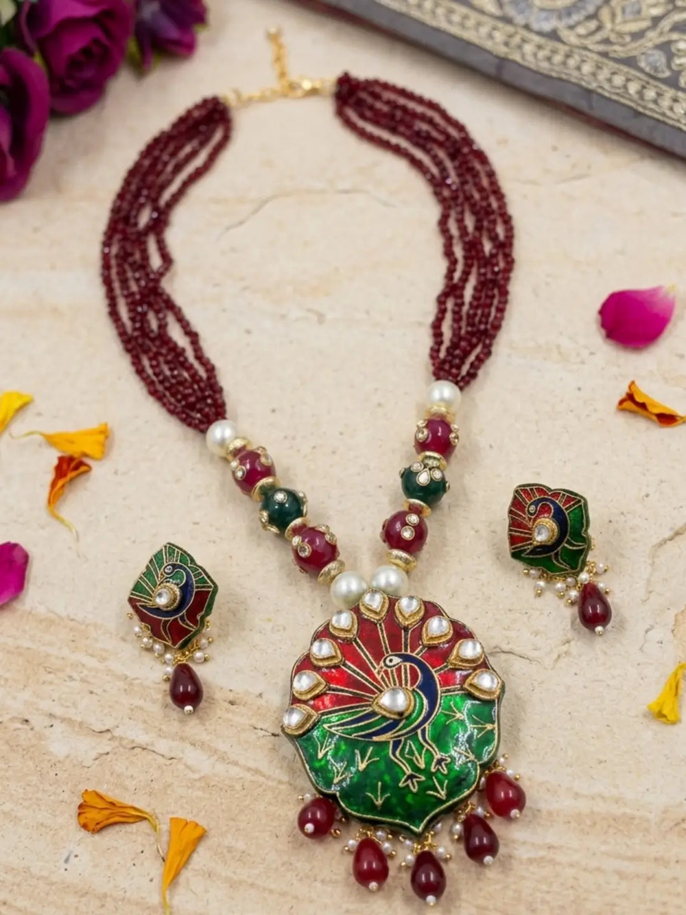 A traditional Indian jewelry set featuring a necklace and matching earrings. The necklace has a large central pendant with a peacock motif in green, red, and gold, accented with white pearls and red beads.