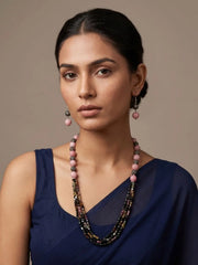 A woman wearing a multi-strand natiral gemstone tourmaline and pink jade beaded necklace with pink and dark metallic beads, matching pink drop earrings, and a navy blue top, set against a solid beige background.