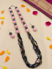 A natural gemtone tourmaline beaded necklace and matching drop earrings displayed on a light beige fabric background with scattered orange and yellow flower petals. The jewelry features round pink beads, dark gray glittery beads, and faceted beads in black, gray, and purple tones.