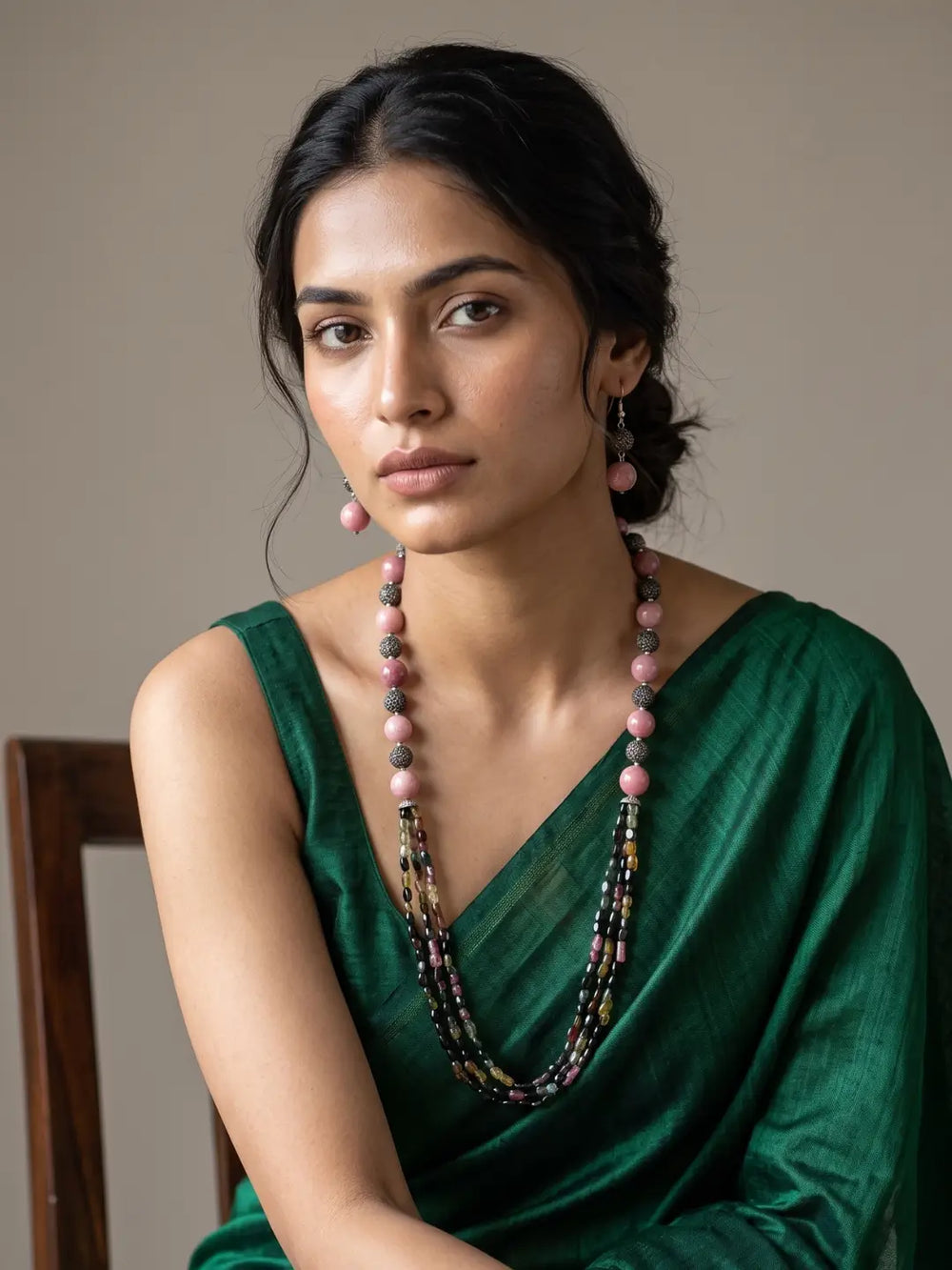 A woman wearing a dark green satin saree and a long multi-strand natural gemstone beaded necklace with pink and dark beads, posed against a neutral beige background.
