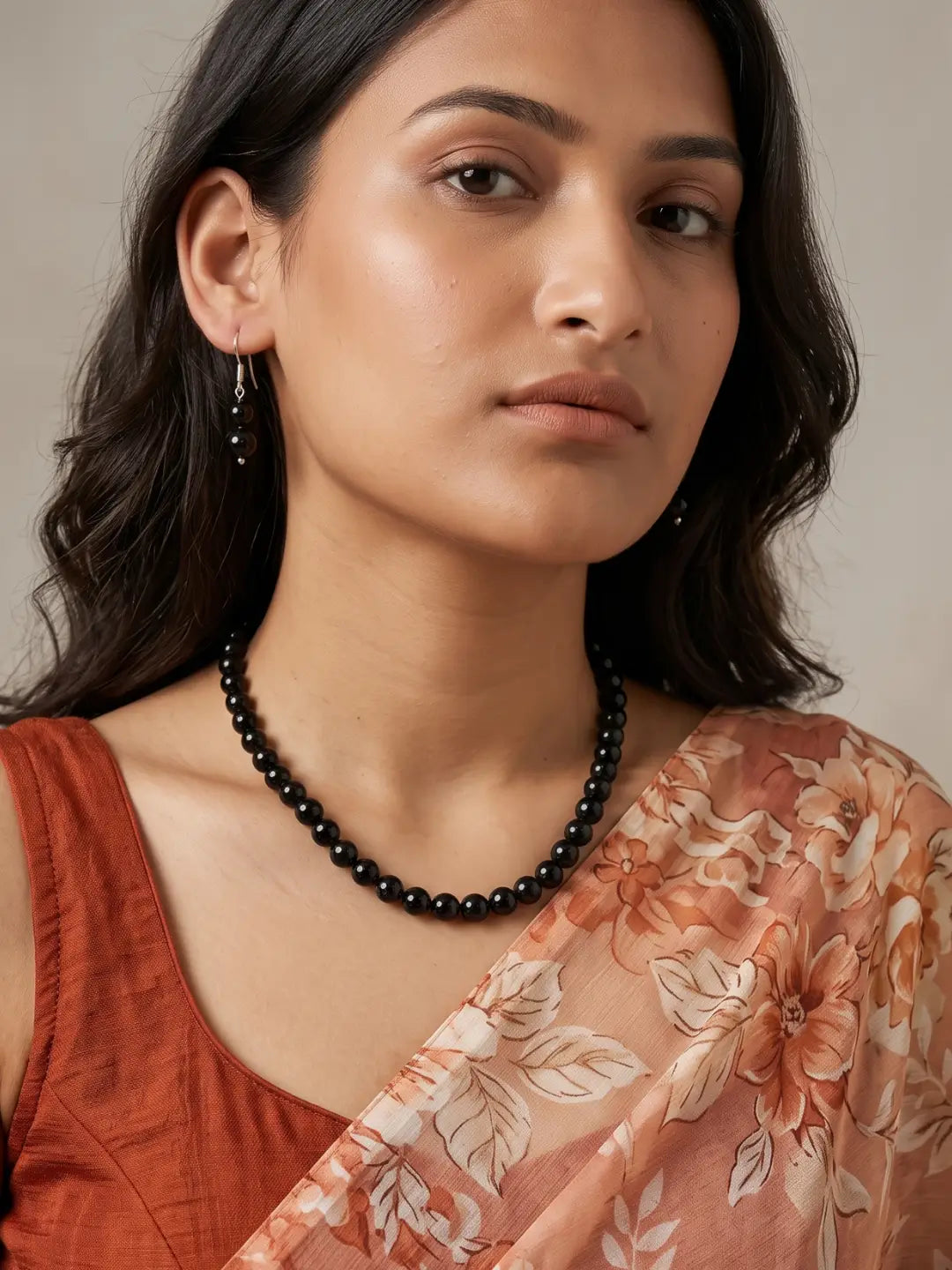 A woman wearing a natural black jade beaded necklace and small black dangling earrings, styled with a rust-colored top and a peach floral dupatta against a neutral beige background.