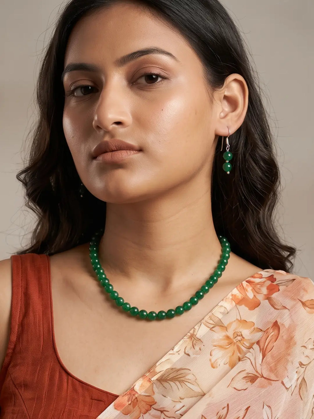 A woman wearing a green beaded necklace and matching green drop earrings, styled with a rust-colored top and a peach floral dupatta against a neutral beige background.