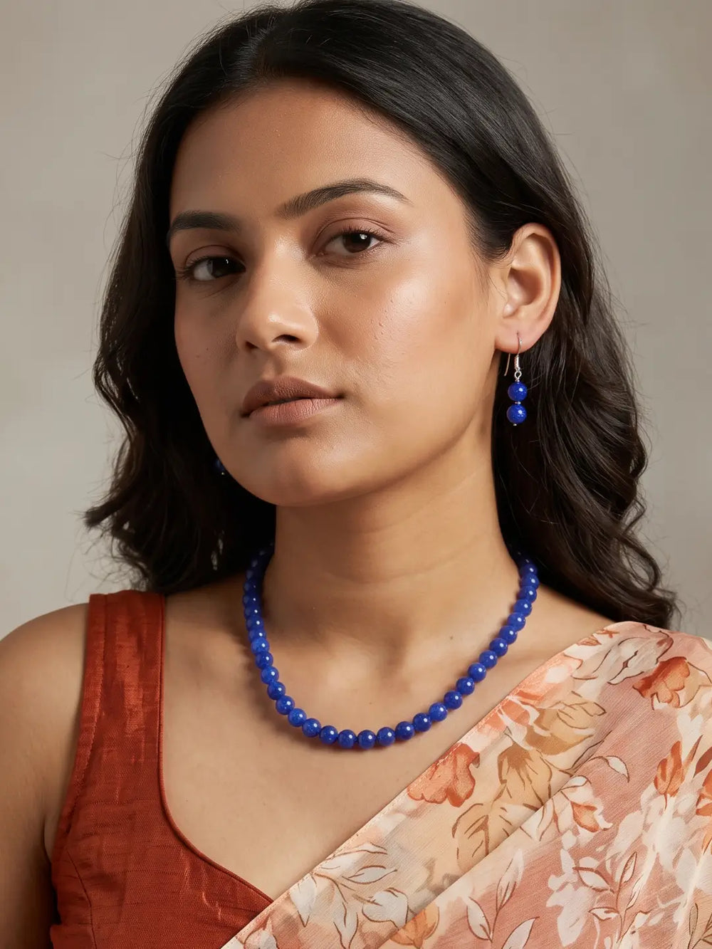A woman wearing a royal blue beaded necklace and matching blue beaded drop earrings, paired with a rust-colored top and a peach floral saree, set against a neutral beige background.