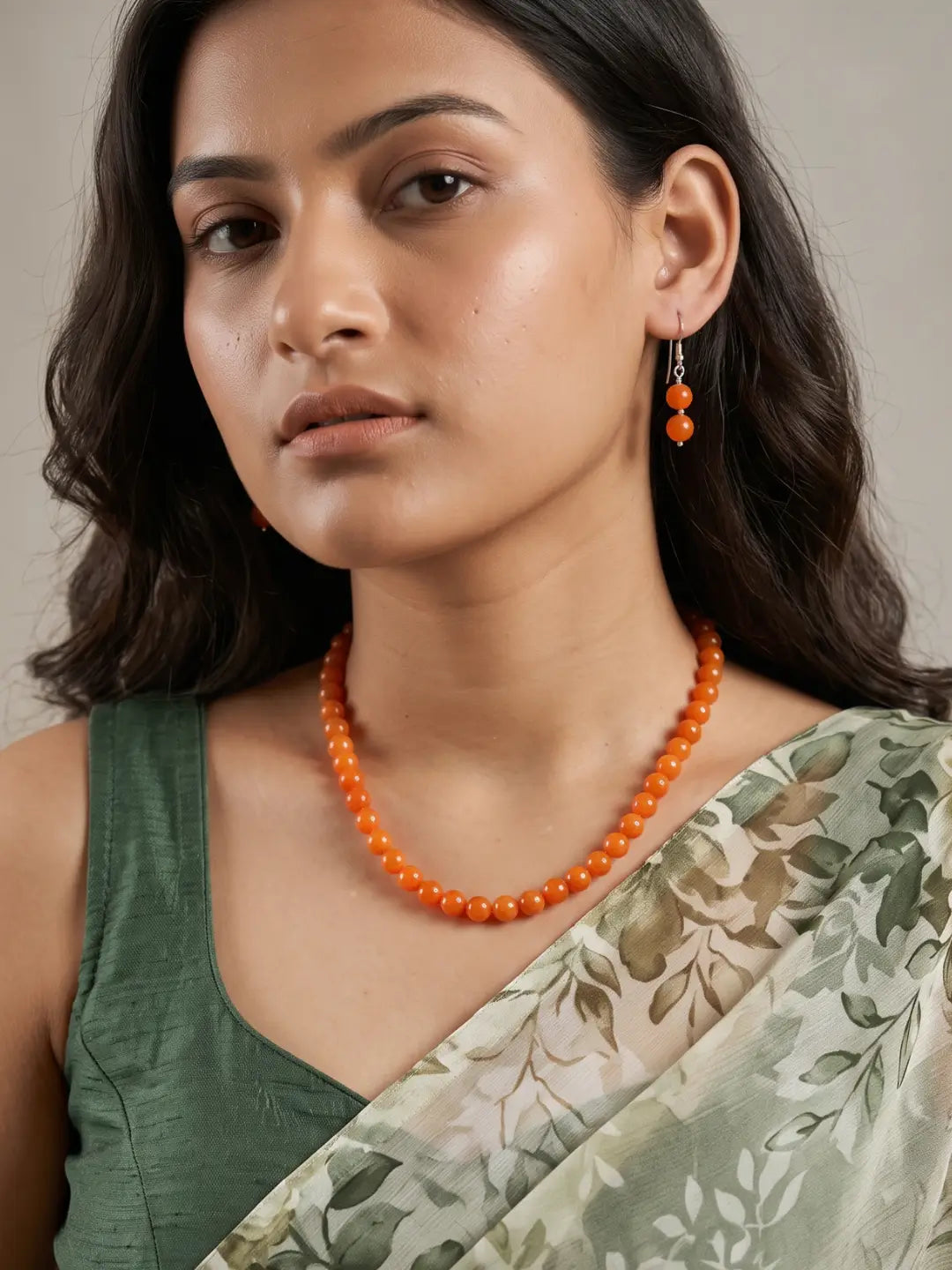 A woman wearing a bright orange beaded necklace and matching orange drop earrings, paired with a green and beige floral printed saree.