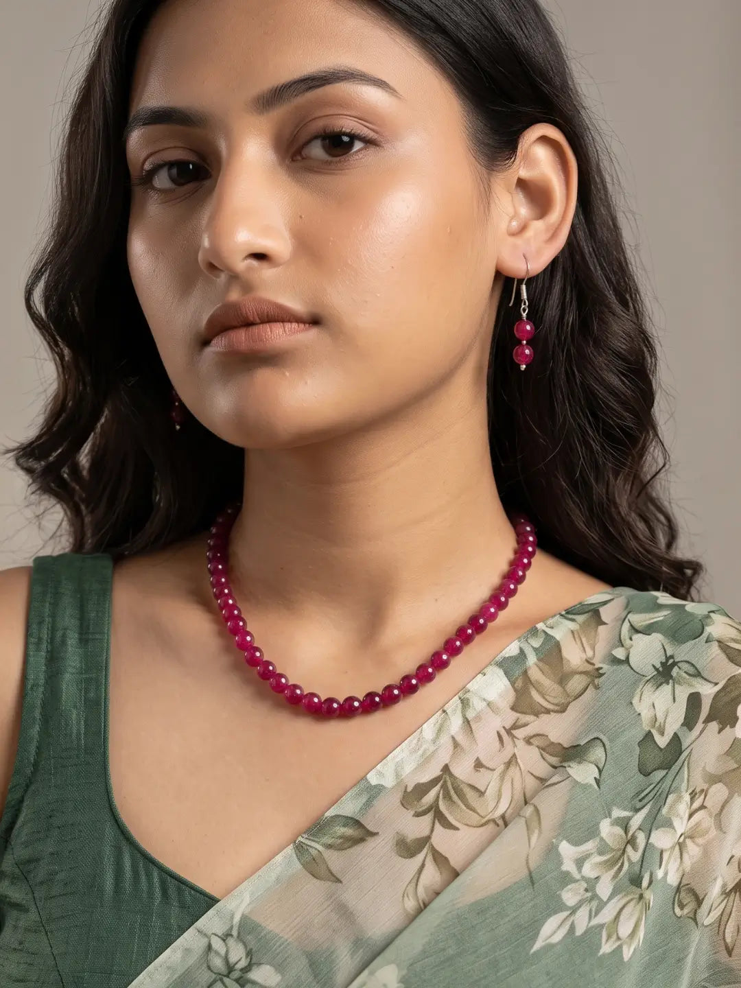 A woman wearing a vibrant semi-precious ruby red jade beaded necklace and matching dangling earrings, paired with a green floral saree.