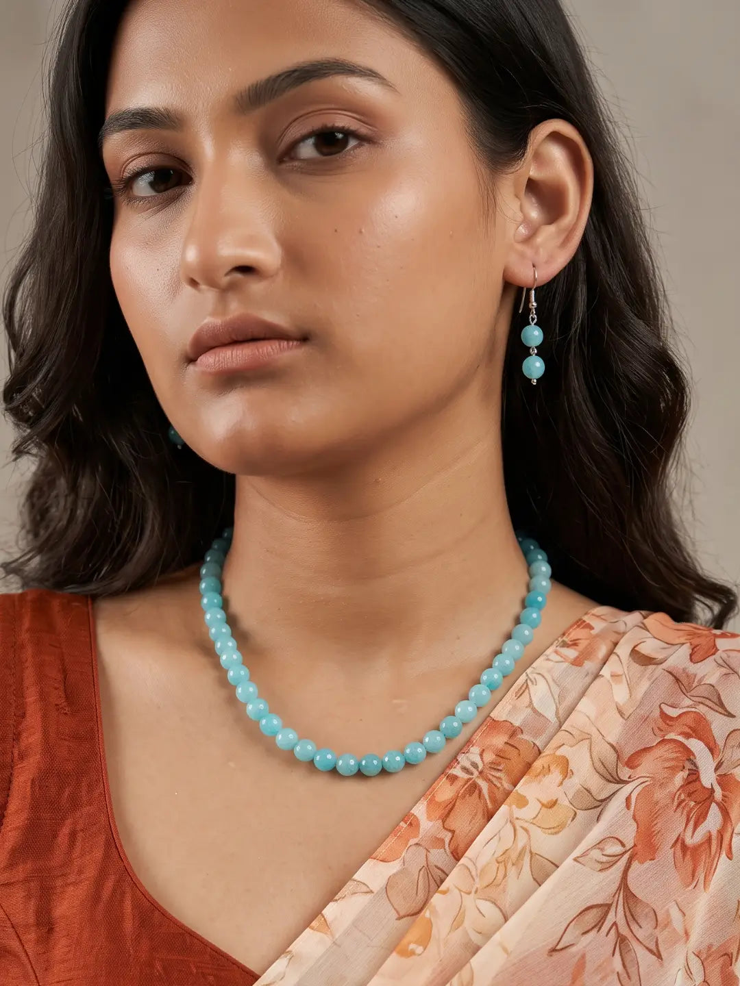 A woman wearing a light blue beaded necklace and matching drop earrings, paired with a rust-colored top and a peach floral sari, set against a neutral beige background.