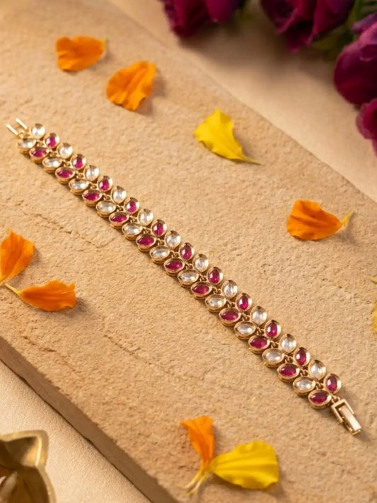 A gold-plated bracelet adorned with rows of clear round stones and pink oval stones, laid diagonally on a beige textured stone slab with scattered orange and yellow flower petals.