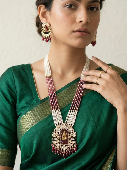 Close view of a woman wearing a traditional red Lakshmi Temple necklace featuring layered pearl strands, an ornate Lakshmi Devi pendant with Kundan Polki detailing and matching earrings.