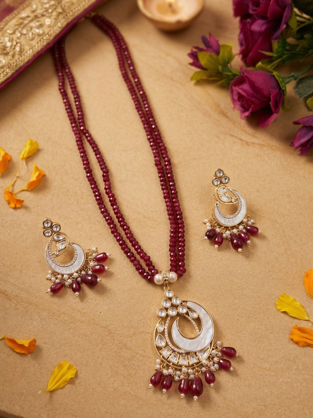 A traditional Indian jewelry set featuring a long beaded necklace and matching earrings. The necklace consists of deep red beads draped over a mother of pearl crescent pendant adorned with white  polki stones and dangling red beads.