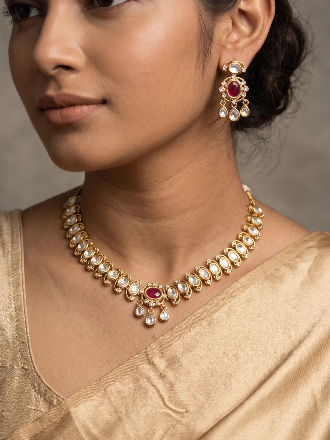 The ruby pink Polki choker necklace set features oval floral motifs, clear CZ and imitation stones, a bold centrepiece, and matching dangler earrings.