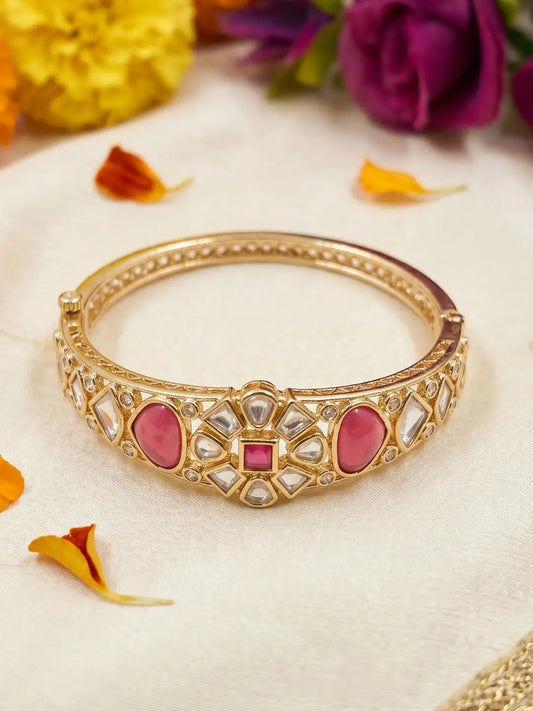 Gold-plated ruby pink Kundan bangle bracelet displayed on a cream fabric with decorative flowers and petals.
