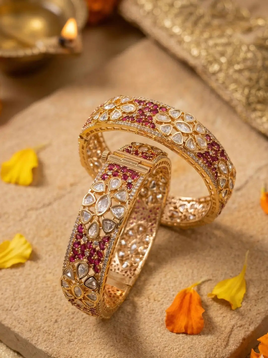 Two ornate gold-plated bangles studded with ruby and polki kundan stones are displayed on a beige stone surface with scattered yellow flower petals.