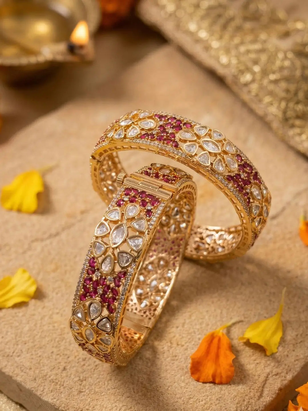 Two ornate gold-plated bangles studded with ruby and polki kundan stones are displayed on a beige stone surface with scattered yellow flower petals.