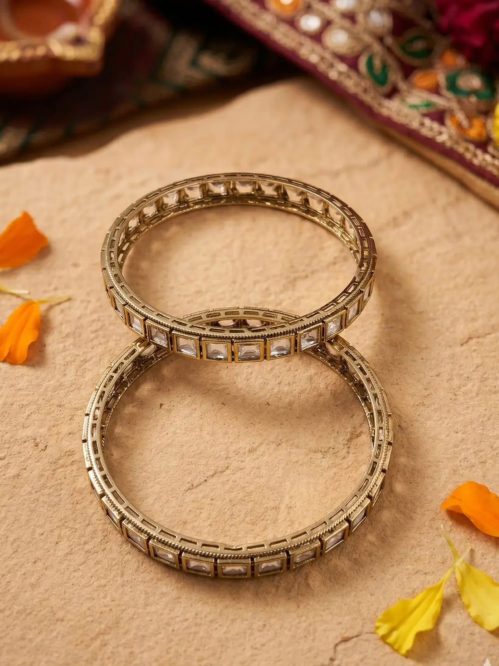 Two victorian plated bangles with clear rectangular polki stones, arranged on a textured beige surface with scattered orange and yellow flower petals.