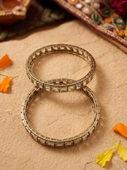 Two victorian plated bangles with clear rectangular polki stones, arranged on a textured beige surface with scattered orange and yellow flower petals.