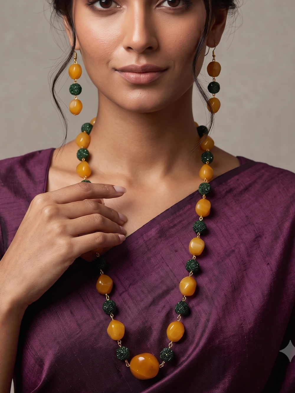 A woman wearing a yellow jade long necklace with green textured beads, a green beaded necklace, styled with a purple sari against a neutral background.