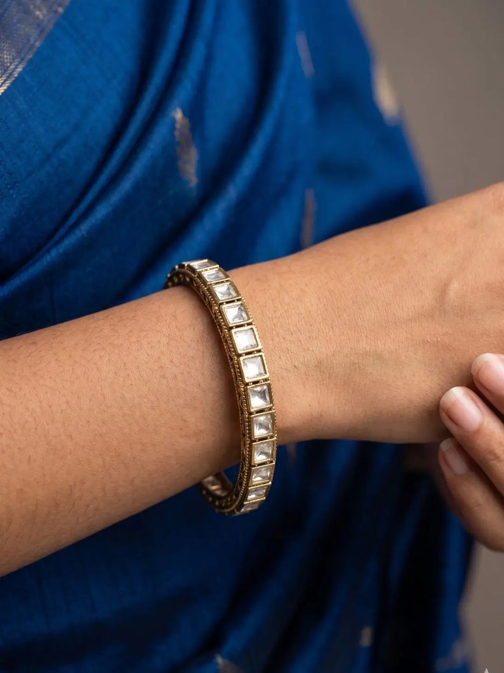 A close-up of a person's wrist wearing a victorian-plated bangle adorned with square white polki stones, set against a blue sari.