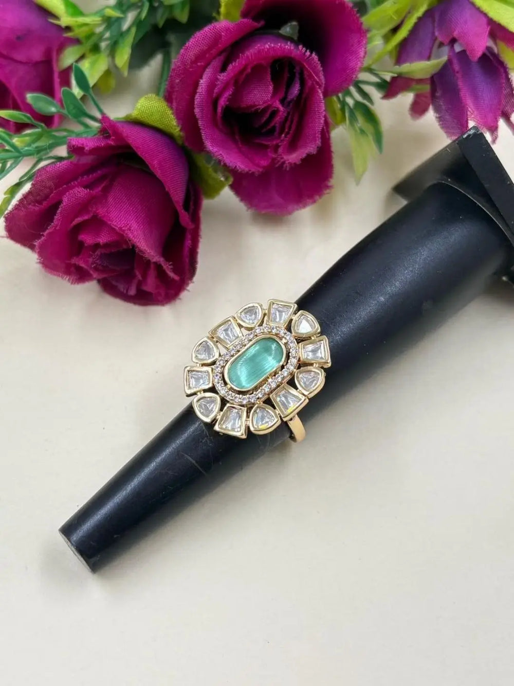 A gold ring with a central mint green oval stone and surrounding clear stones, displayed on a black ring stand with magenta roses in the background.