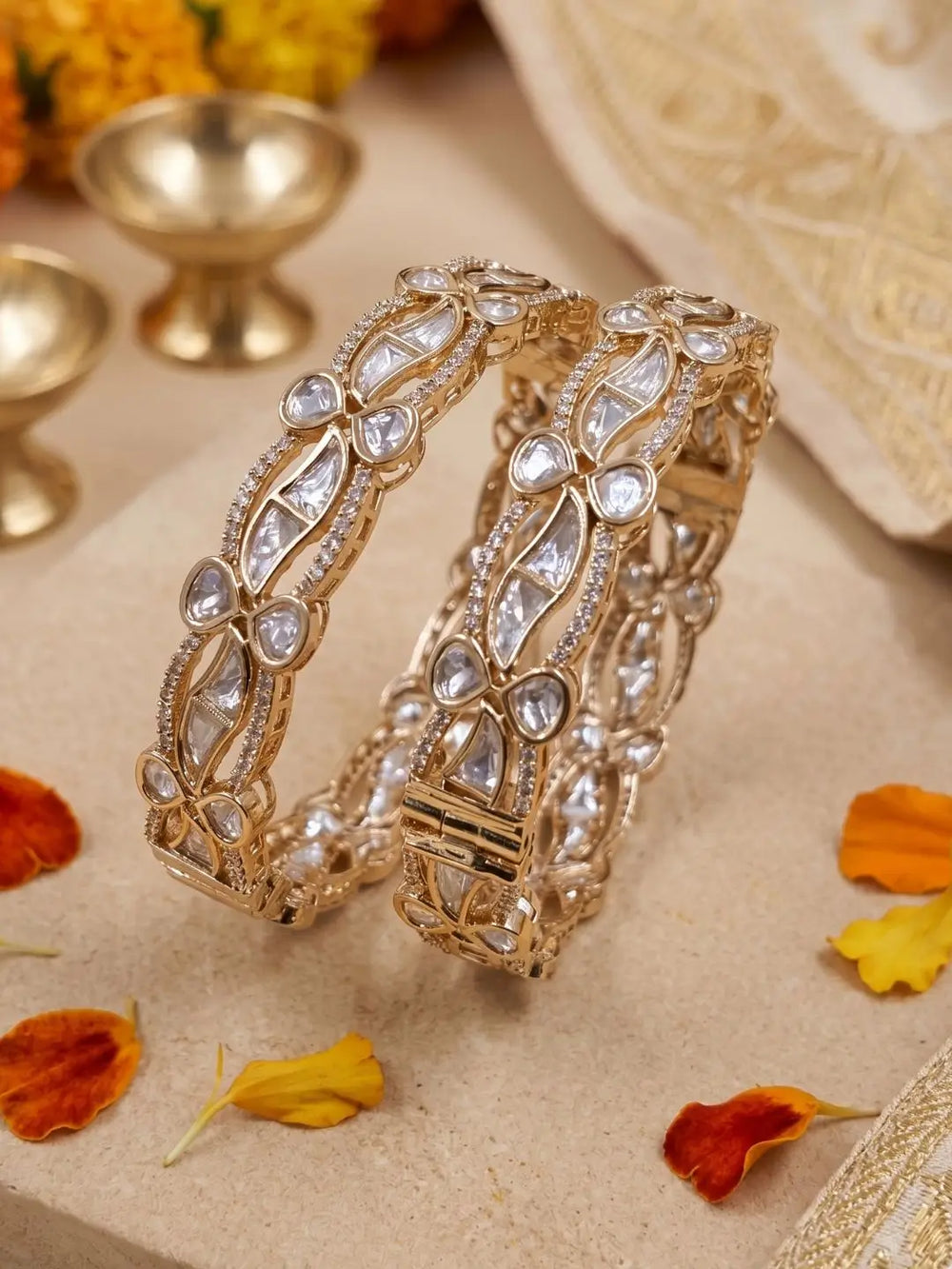 A pair of ornate gold-toned bangles decorated with clear stones, displayed on a beige textured surface with scattered orange and yellow flower petals and blurred decorative props in the background.