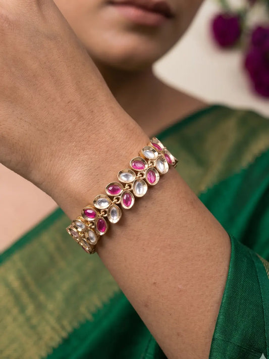 A close-up of a woman's wrist wearing a gold-plated bracelet adorned with pink and clear teardrop-shaped stones, set against a green saree.