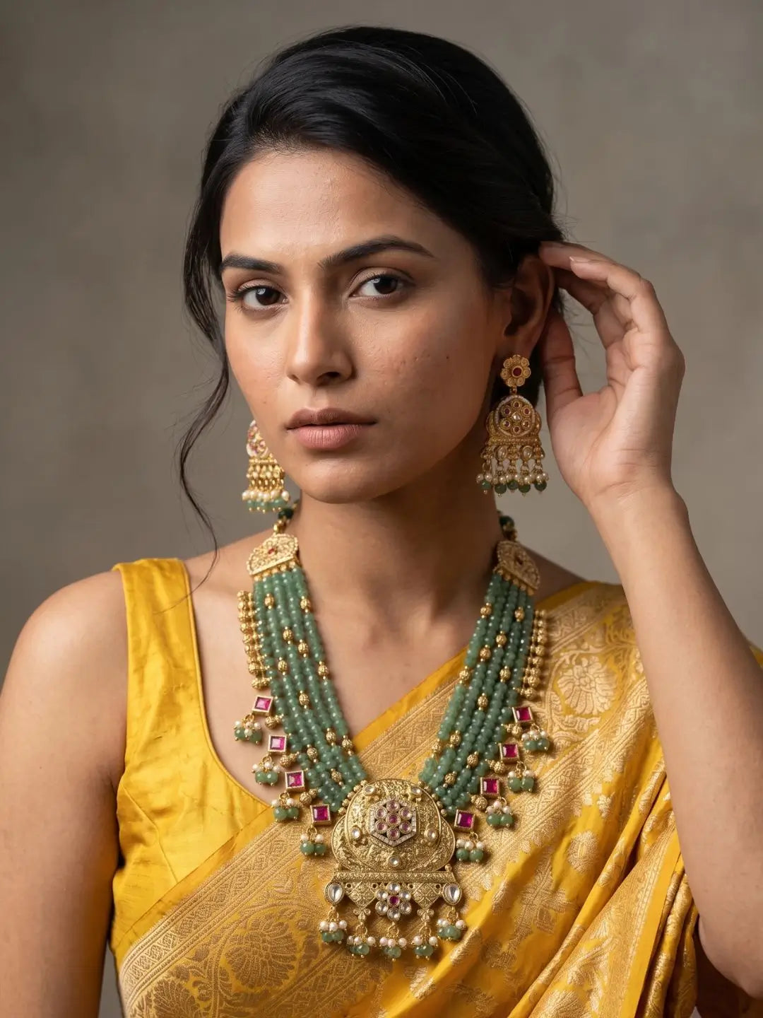A woman wearing a traditional Indian yellow saree and a large green-and-gold statement necklace with matching earrings, posed against a neutral gray background.