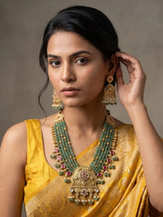 A woman wearing a traditional Indian yellow saree and a large green-and-gold statement necklace with matching earrings, posed against a neutral gray background.