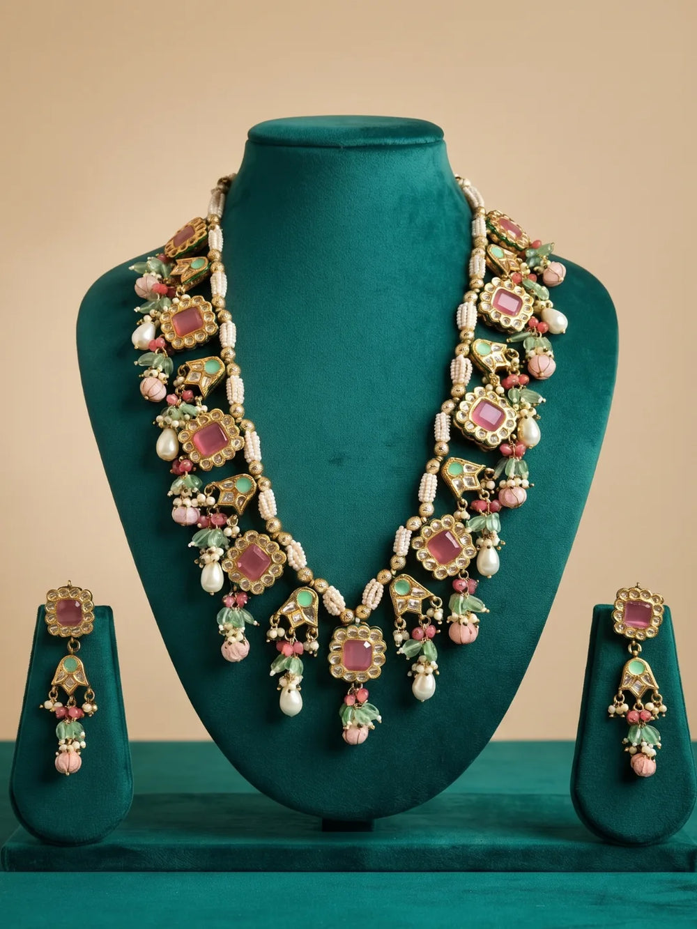 An ornate gold jewelry set displayed on a dark teal velvet mannequin bust, including a multi-colour necklace and matching drop earrings. The jewelry features pink jadau stones, green enamel details, and white pearl-like beads, set against a solid beige background.
