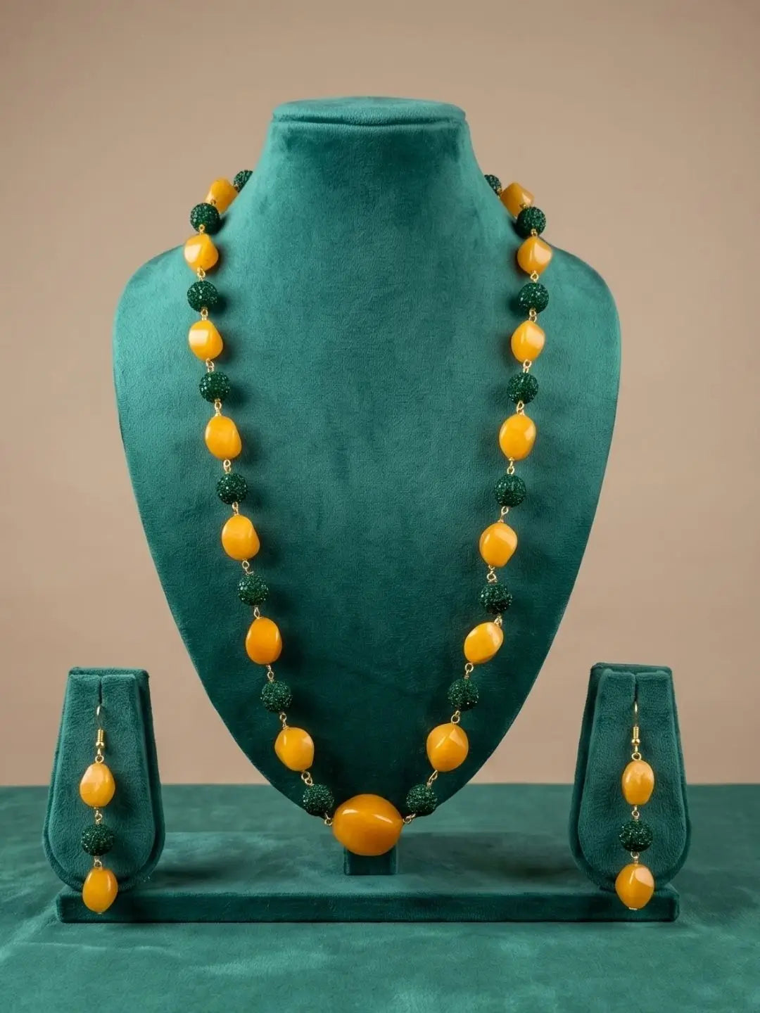 A jewelry set displayed on a teal velvet mannequin bust, featuring a long beaded necklace and matching drop earrings. The beads alternate between smooth, rounded golden-yellow beads and textured dark green beads, with small gold spacers.