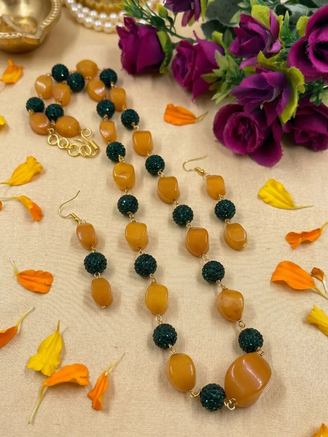 A beaded necklace and matching drop earrings displayed on a beige fabric surface with scattered orange and yellow flower petals. The jewelry features alternating smooth yellow jade beads and dark green textured round beads, with small gold-tone links and hooks.