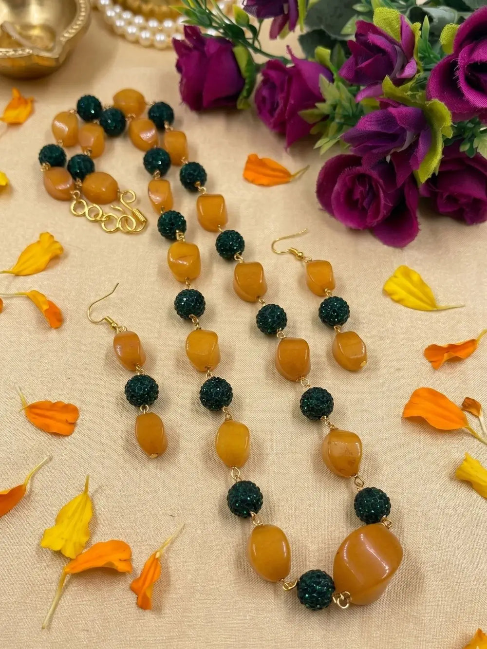 A beaded necklace and matching drop earrings displayed on a beige fabric surface with scattered orange and yellow flower petals. The jewelry features alternating smooth yellow jade beads and dark green textured round beads, with small gold-tone links and hooks.
