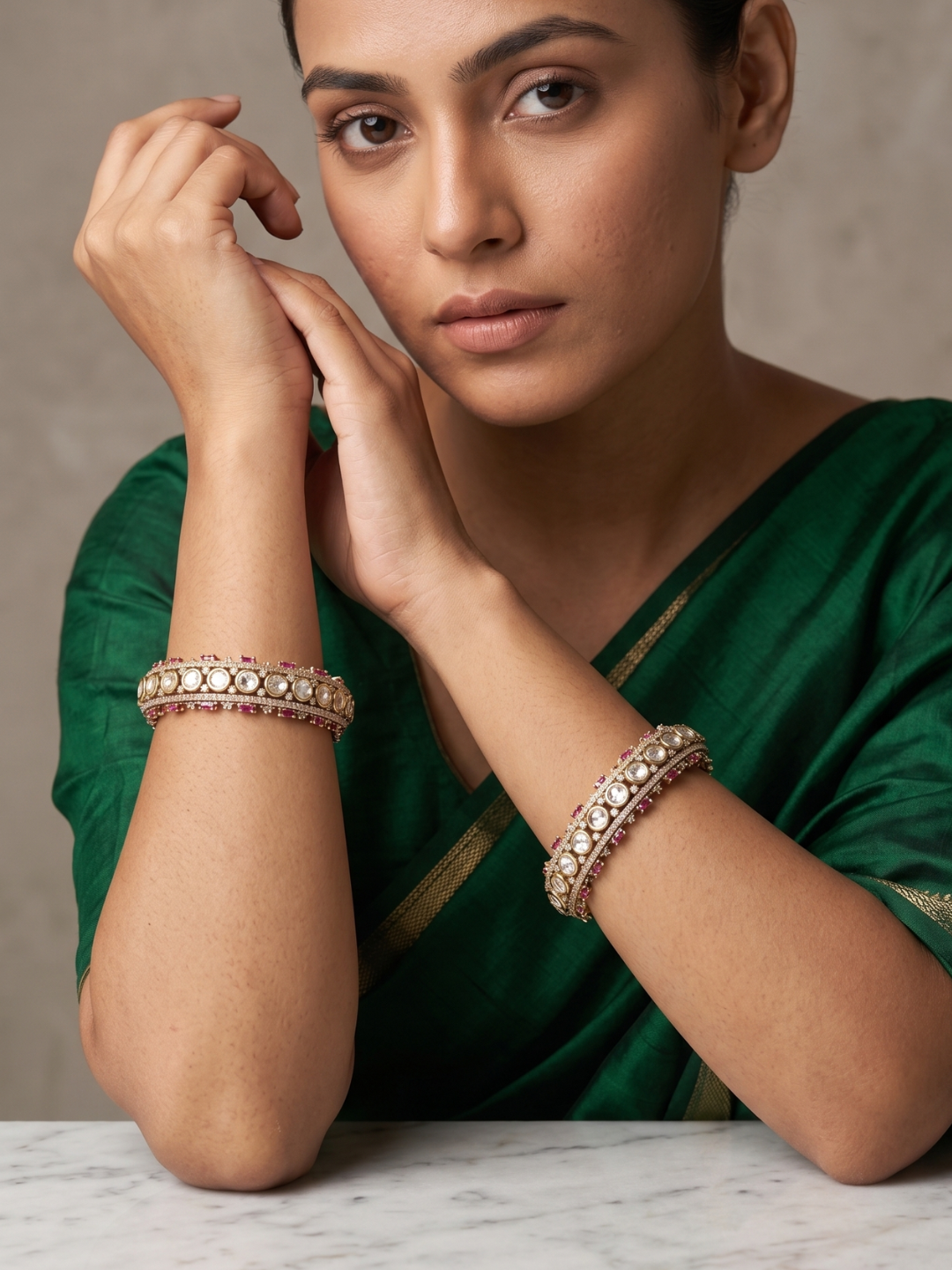 A woman wearing a dark green sari and matching green blouse, showcasing two ornate gold-toned bangles adorned with clear stones on her wrists, set against a neutral gray background.