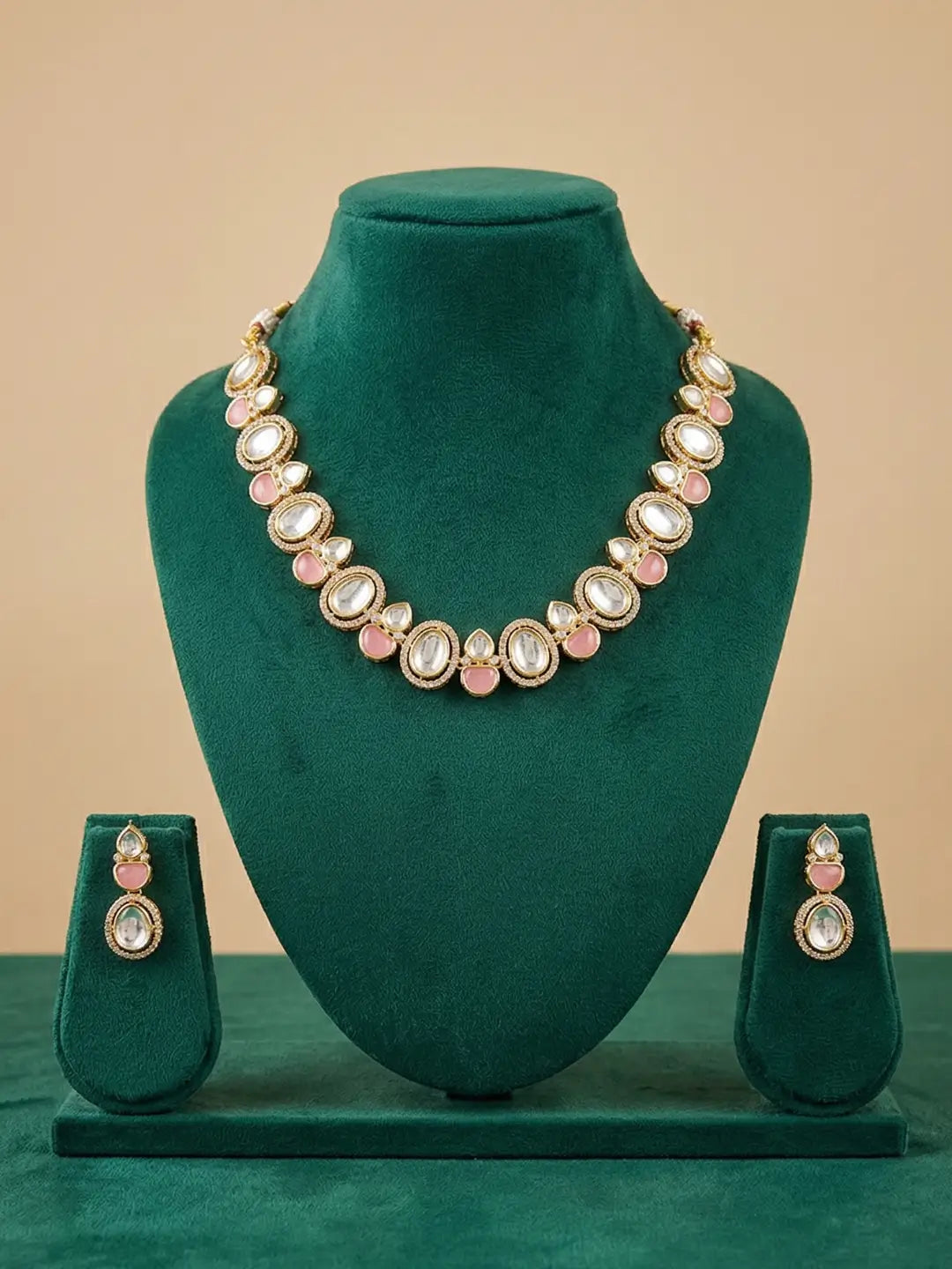 Gold-plated jewelry set featuring a necklace and matching earrings with pink stones and white stones, displayed on a dark green velvet jewelry stand against a beige background.
