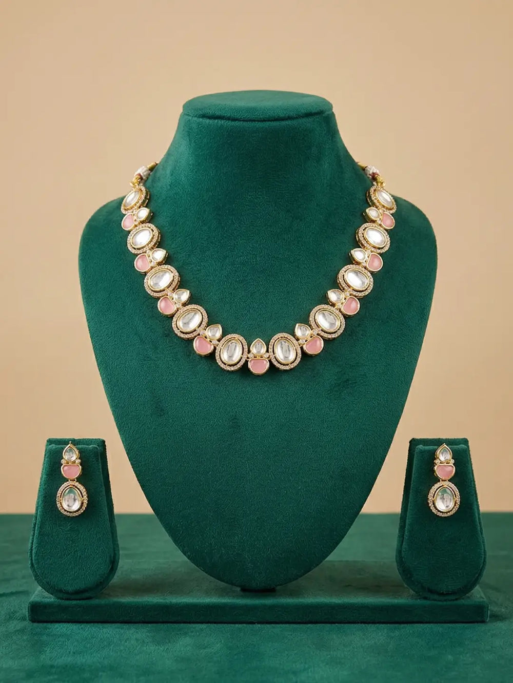 Gold-plated jewelry set featuring a necklace and matching earrings with pink stones and white stones, displayed on a dark green velvet jewelry stand against a beige background.