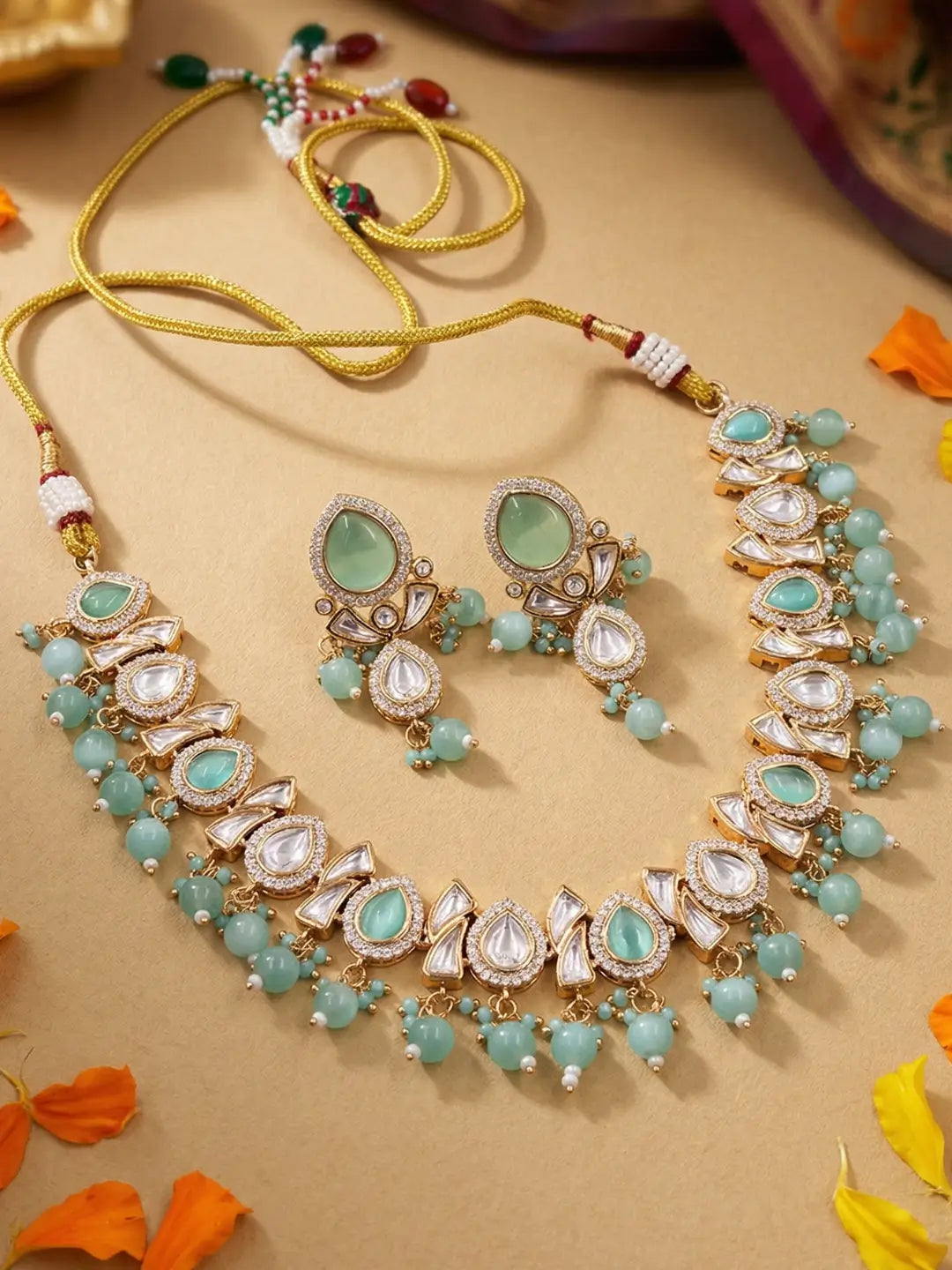 A traditional Indian jewelry set featuring a statement necklace and matching earrings. The set is embellished with light green stones, clear polki crystals, and small green beads on a gold-toned chain, displayed on a beige background with decorative orange leaves.