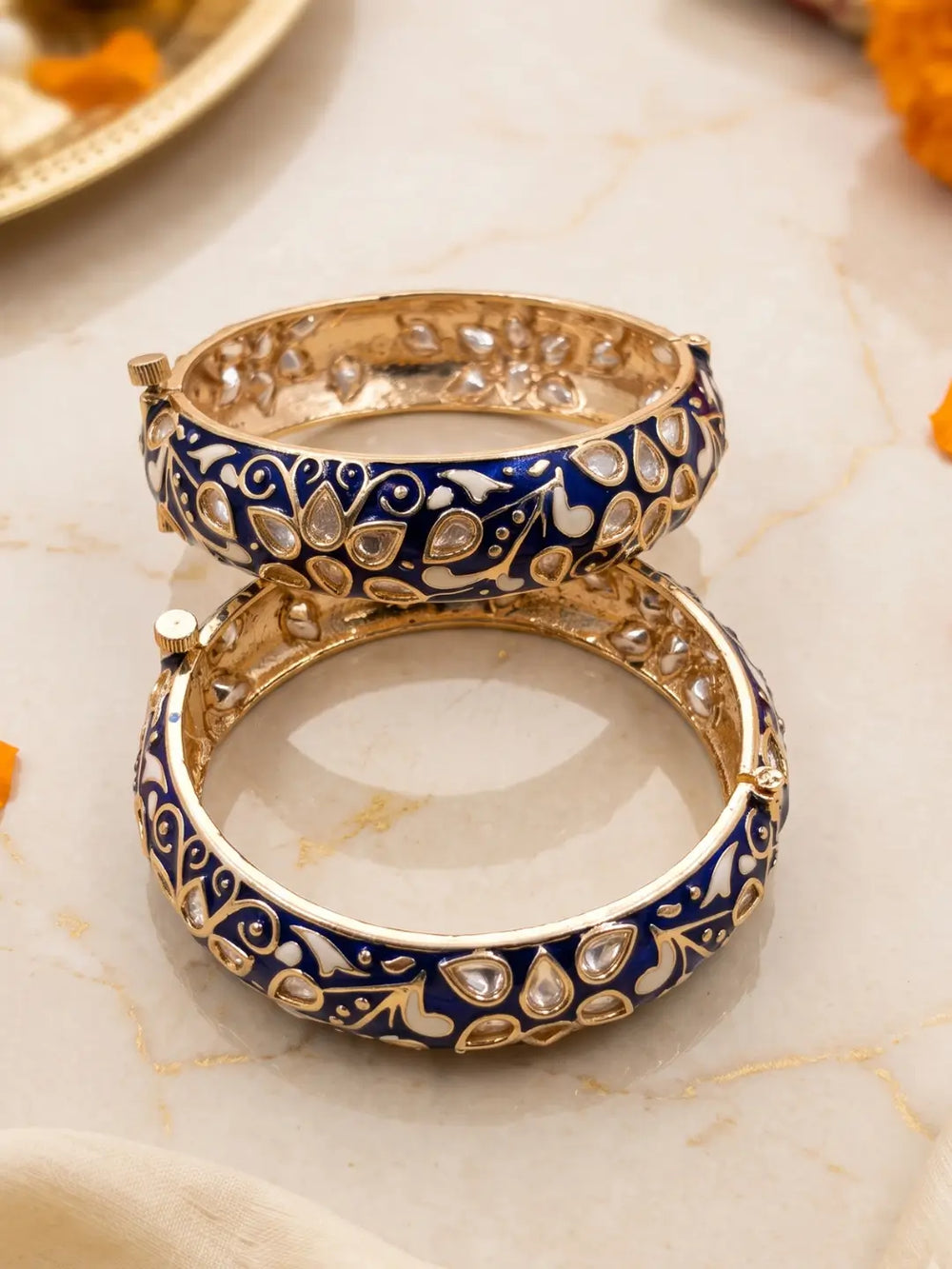 Two stacked Indian-style blue enamel bangles with a deep blue enamel body and intricate gold-toned floral patterns, displayed on a light marble surface.
