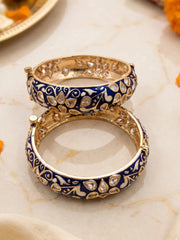 Two stacked Indian-style blue enamel bangles with a deep blue enamel body and intricate gold-toned floral patterns, displayed on a light marble surface.