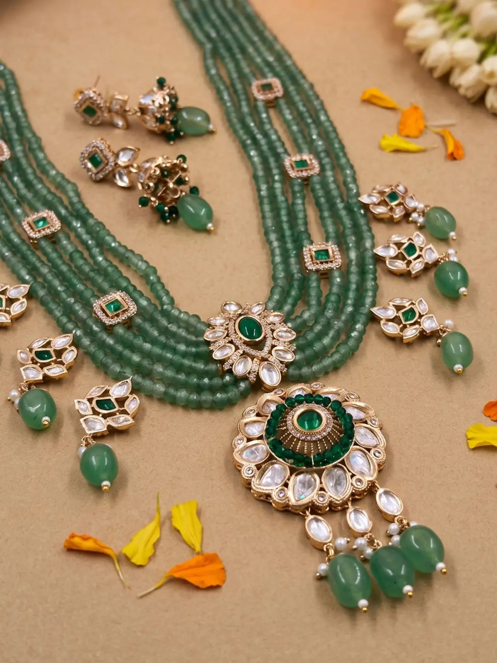 A traditional Indian kundan jewelry set featuring a multi-strand green beaded necklace with a large green and gold pendant, matching earrings arranged on a beige surface with decorative flower petals.