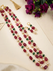 A gold-plated beaded jewelry set featuring a dual-strand necklace and matching earrings with red beads, white pearls, and green gemstone accents, displayed on a cream-colored surface with decorative purple roses and a gold tassel.