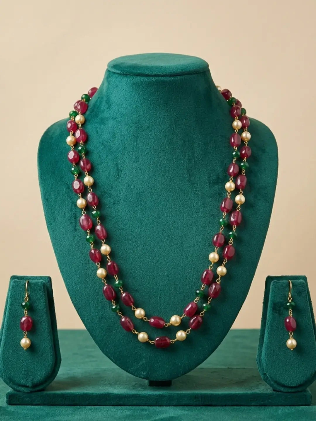 A two-strand beaded necklace and matching drop earrings displayed on a dark green velvet jewelry bust. The necklace features alternating strands of deep red faceted beads, cream-colored pearl-like beads, and small green spacer beads, with gold-toned metal findings.